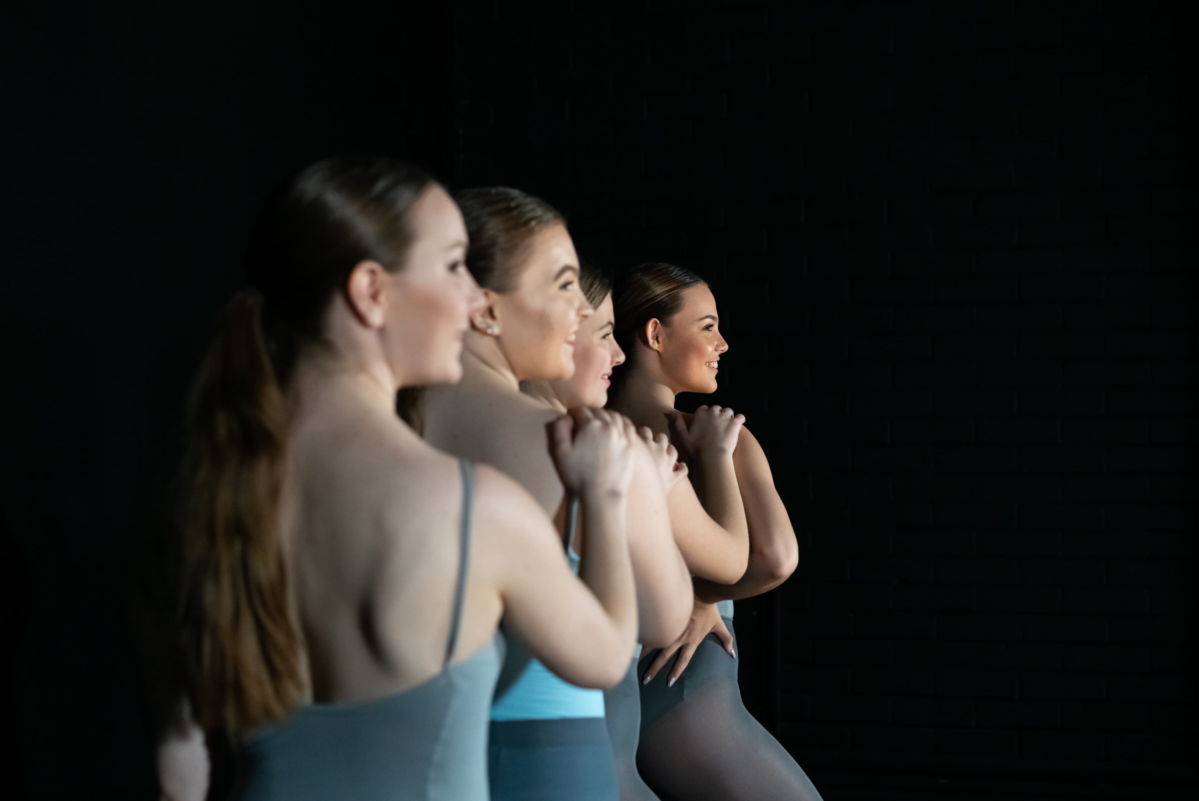 Four dancers stand in a diagonal line with a hand on the shoulder of the person in front.