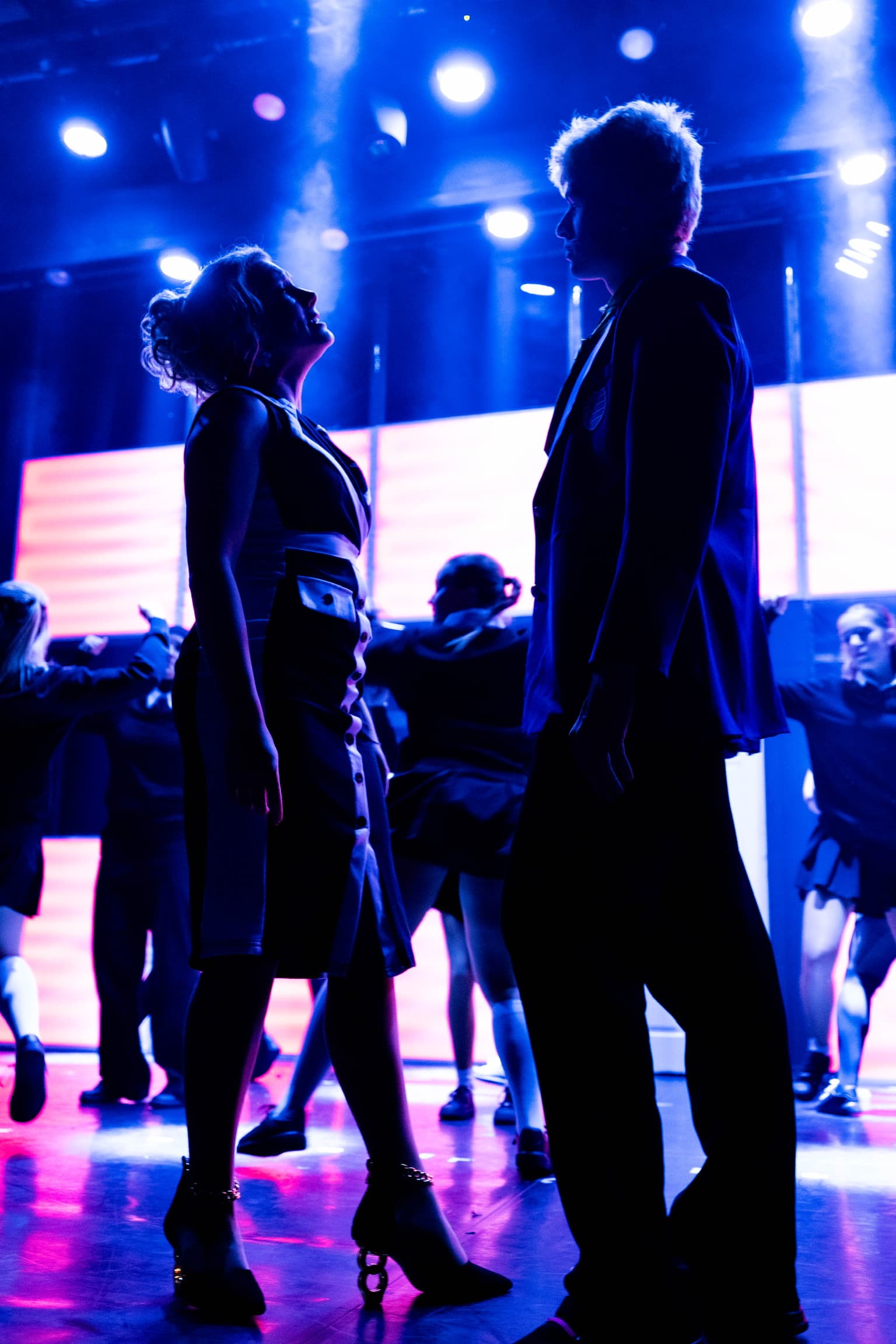 Miss Hedge and Jamie stand silhouetted at the front of the stage. They are lit from behind by bright white light. The LED wall is lit up red.