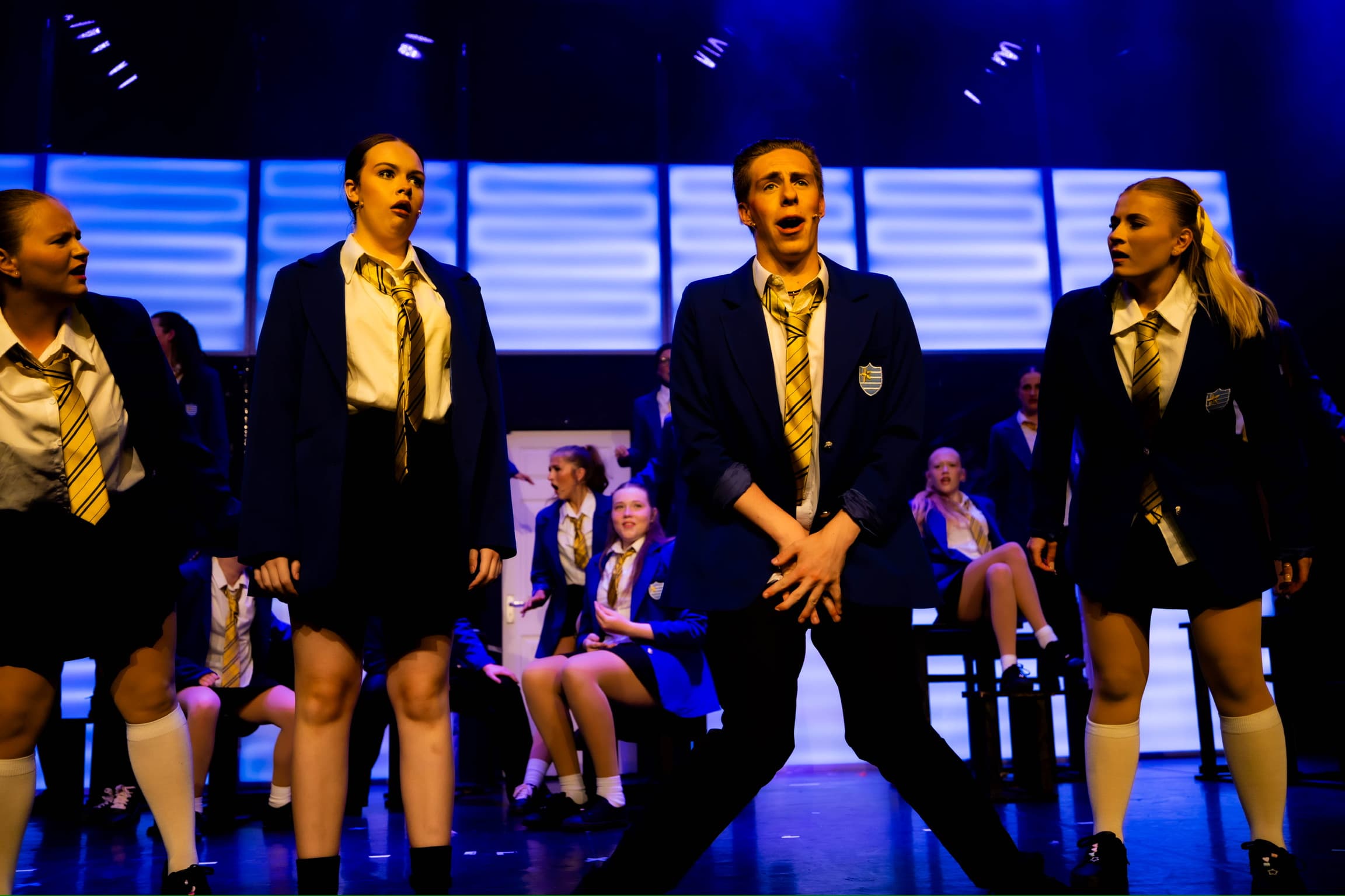 A student dances at the front of the stage, holding his hands in front of his waist. Other classmates look at him. The stage and LED wall are lit up blue.