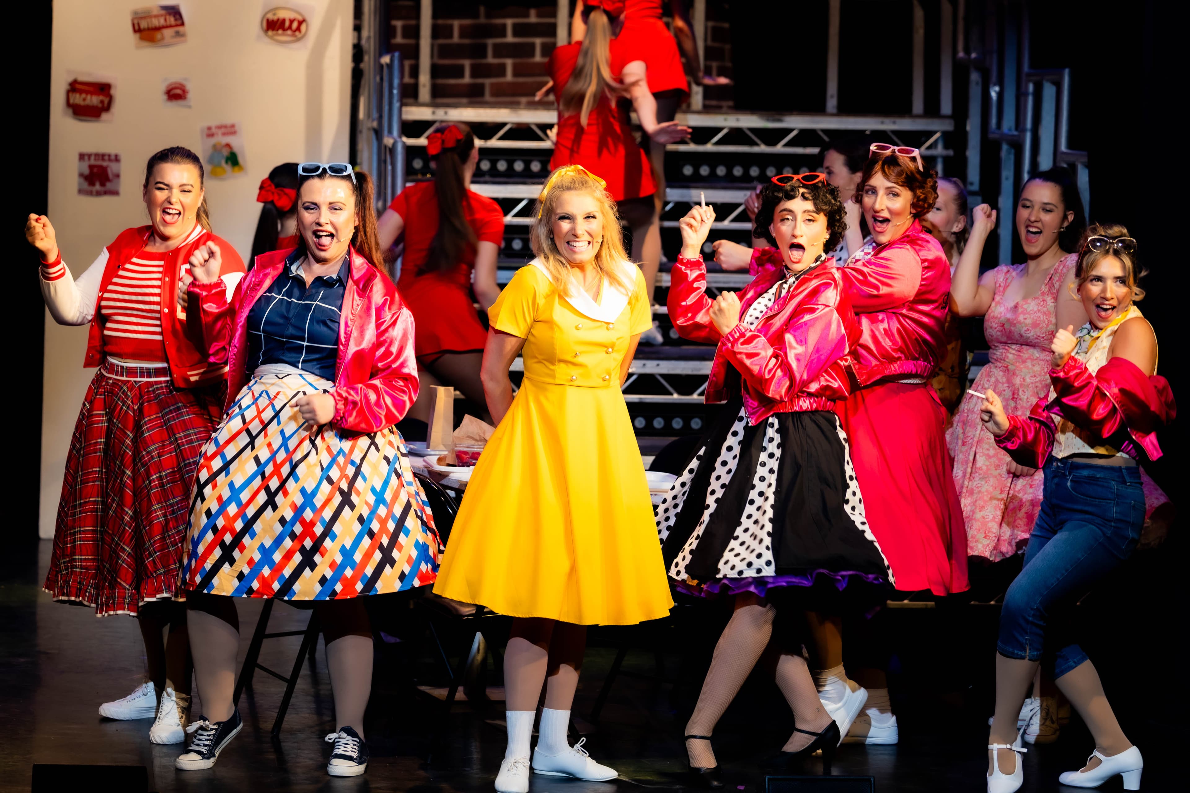 Sandy stands smiling in a yellow dress on stage while the Pink Ladies dance around her.