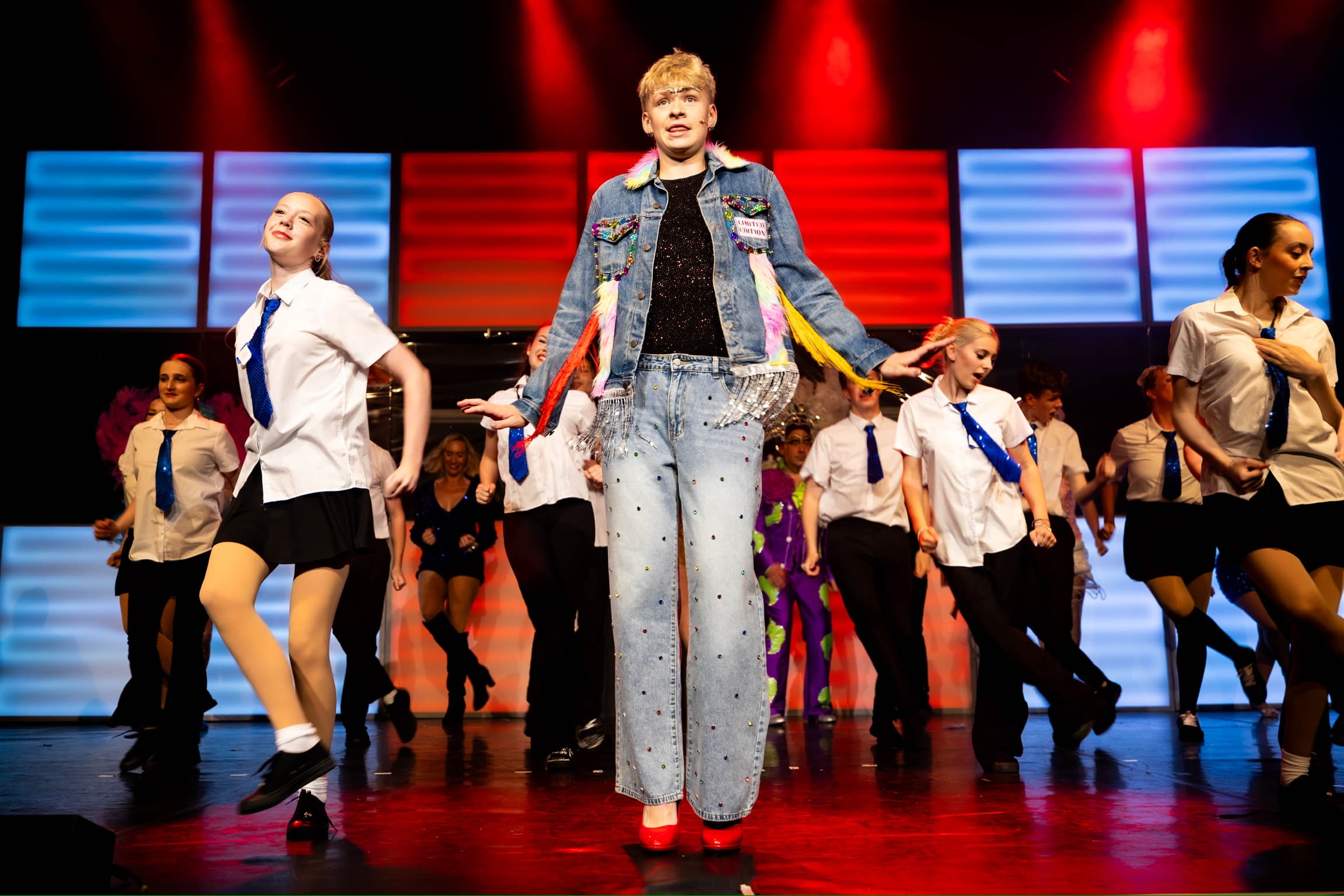 Jamie stands at the front of the stage in full denim and his heels. He is surrounded by dancers in shirts and sparkly blue ties. The LED wall is lit up red and white.