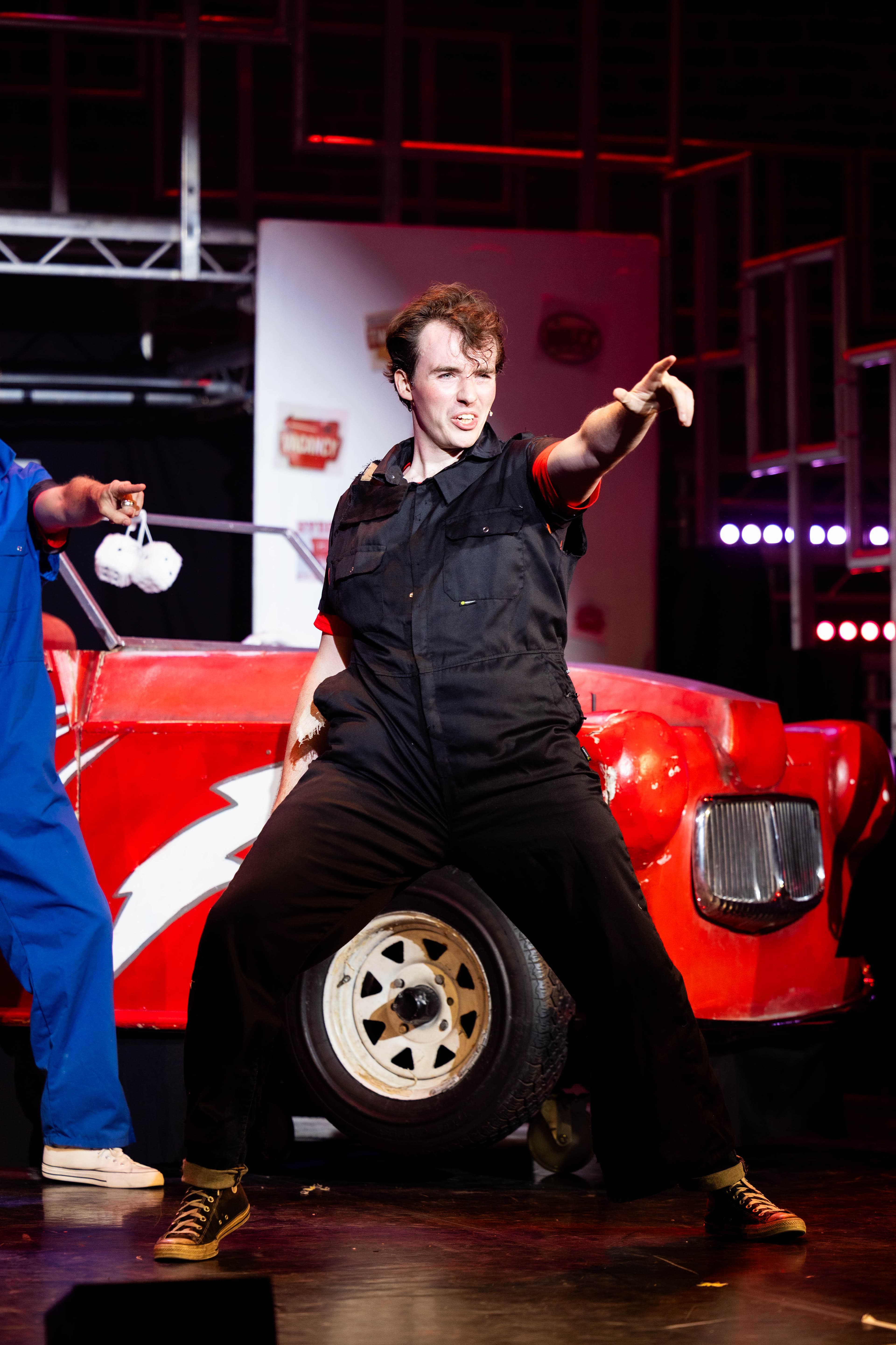 Kenickie dances in a black mechanic boiler suit in front of Greased Lighting. He has his arm out pointing at the audience.