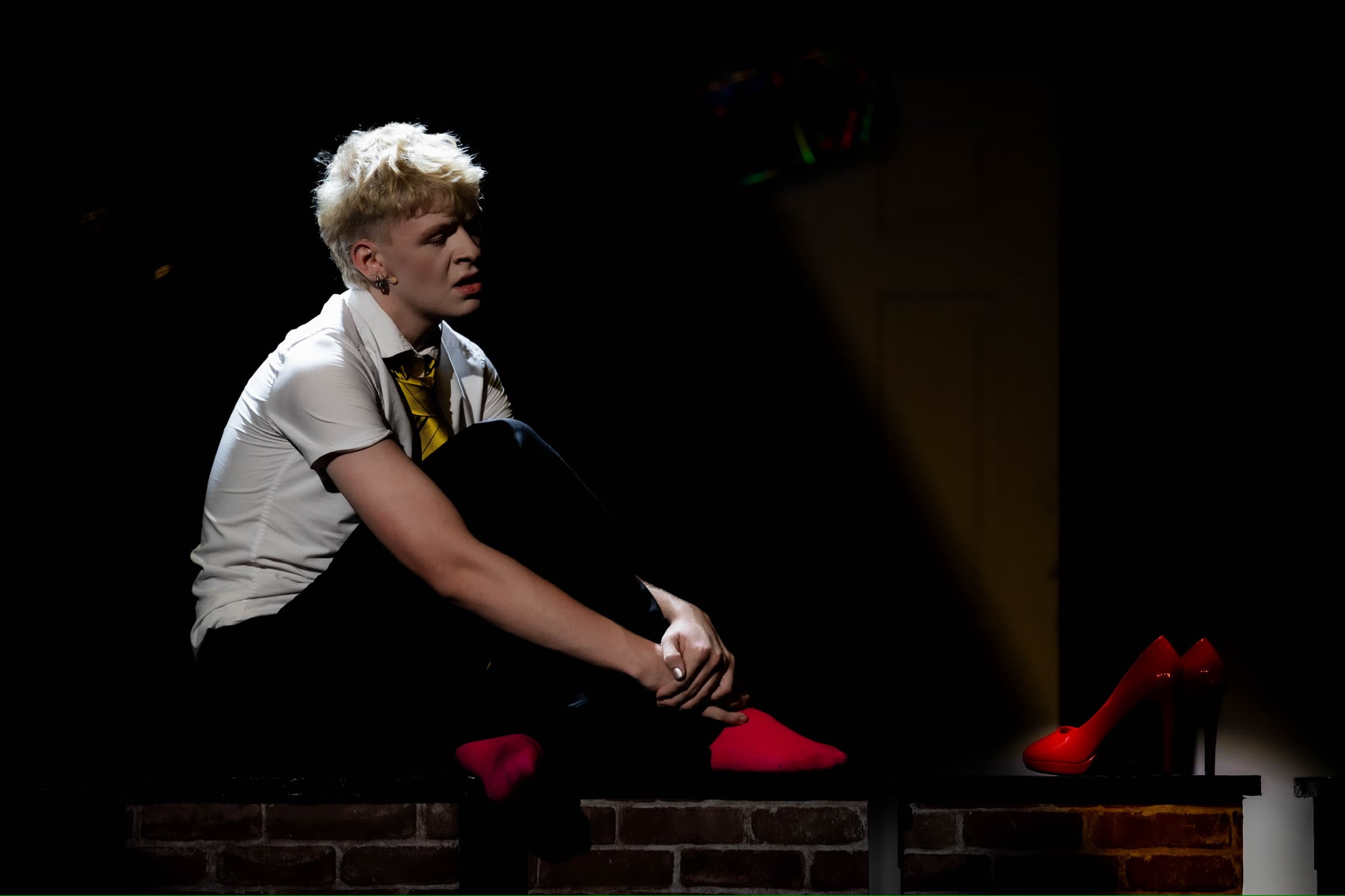 Jamie sits on a wall and looks at his red high heels while singing. The stage is dark and he is light in white.