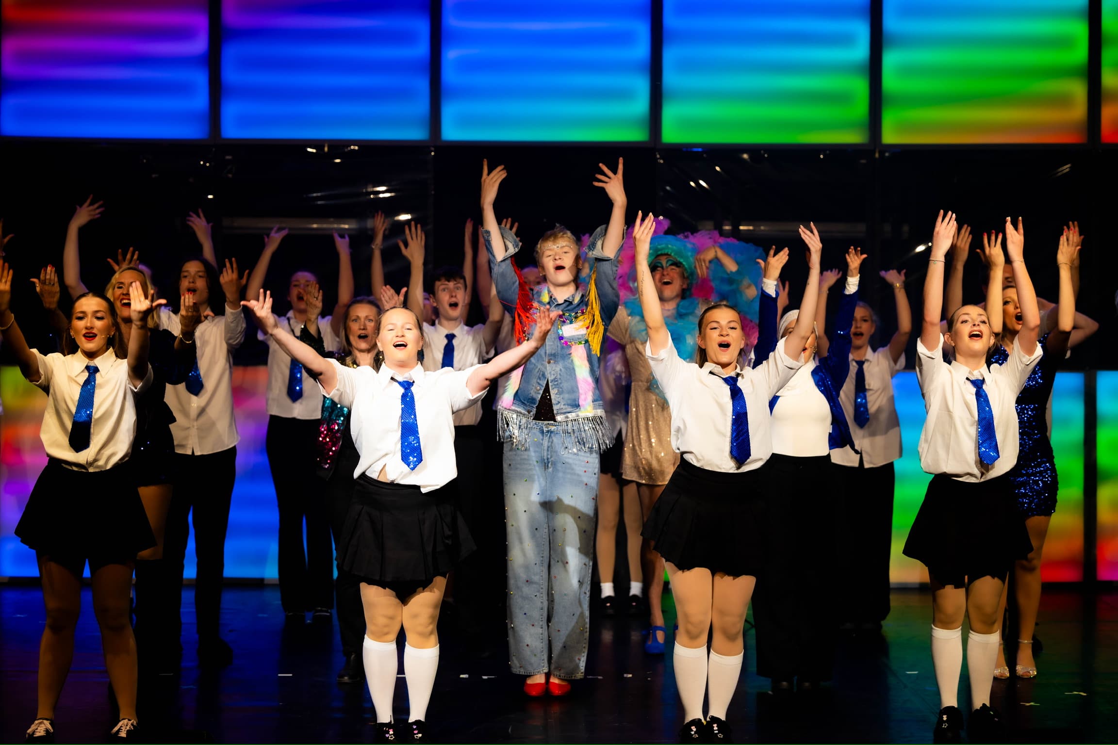 The cast of Jamie pose during the finale. Jamie stands in the middle surrounded by students in shirts and sparkly blue ties. The LED wall is lit up in a rainbow.