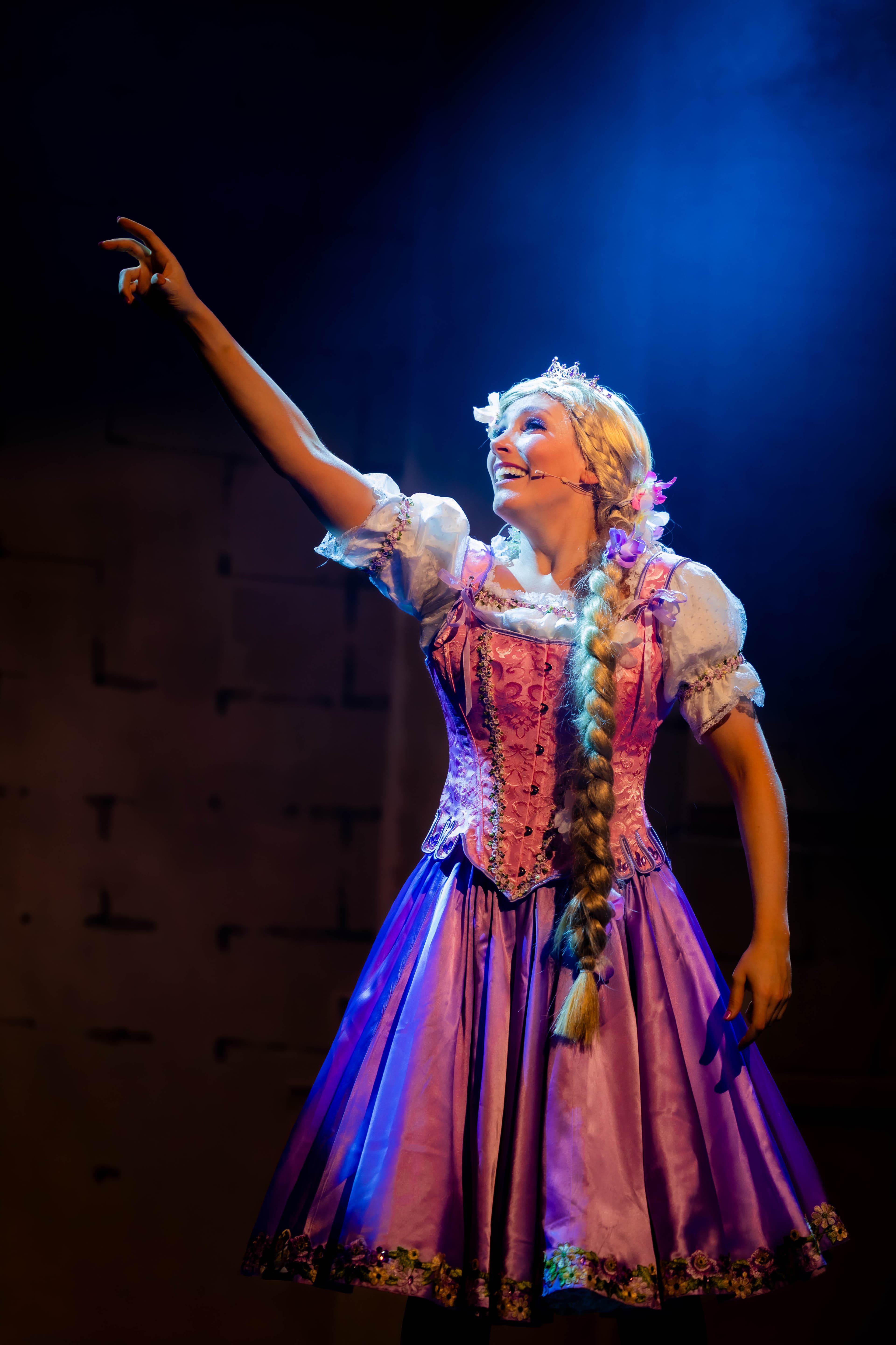 Rapunzel poses pointing into the air while singing. She is backlit from above by a soft blue spotlight.