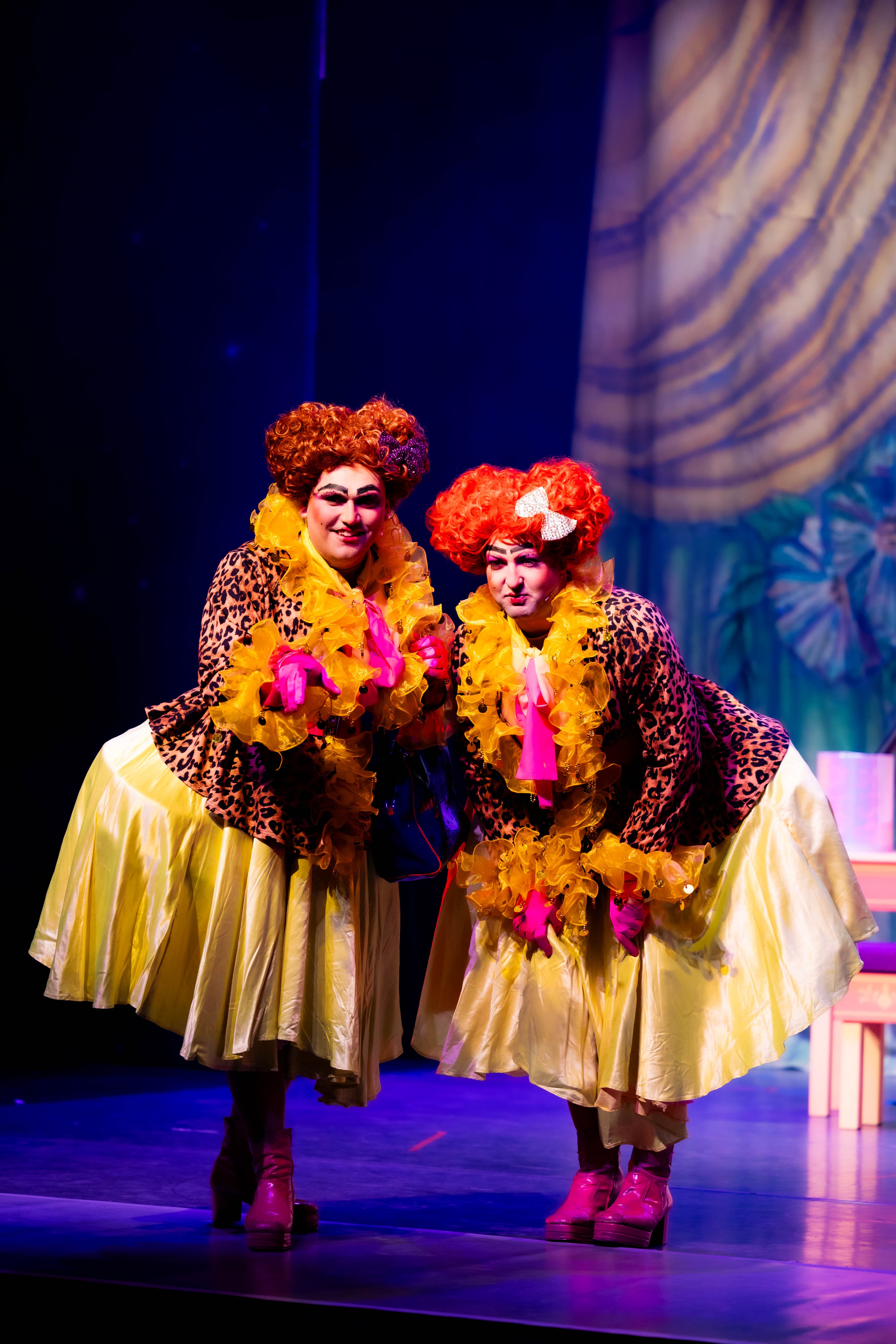Lenore and Zoflora bend down to interact with the audience. They wear matching leopard print dresses with yellow skirts.