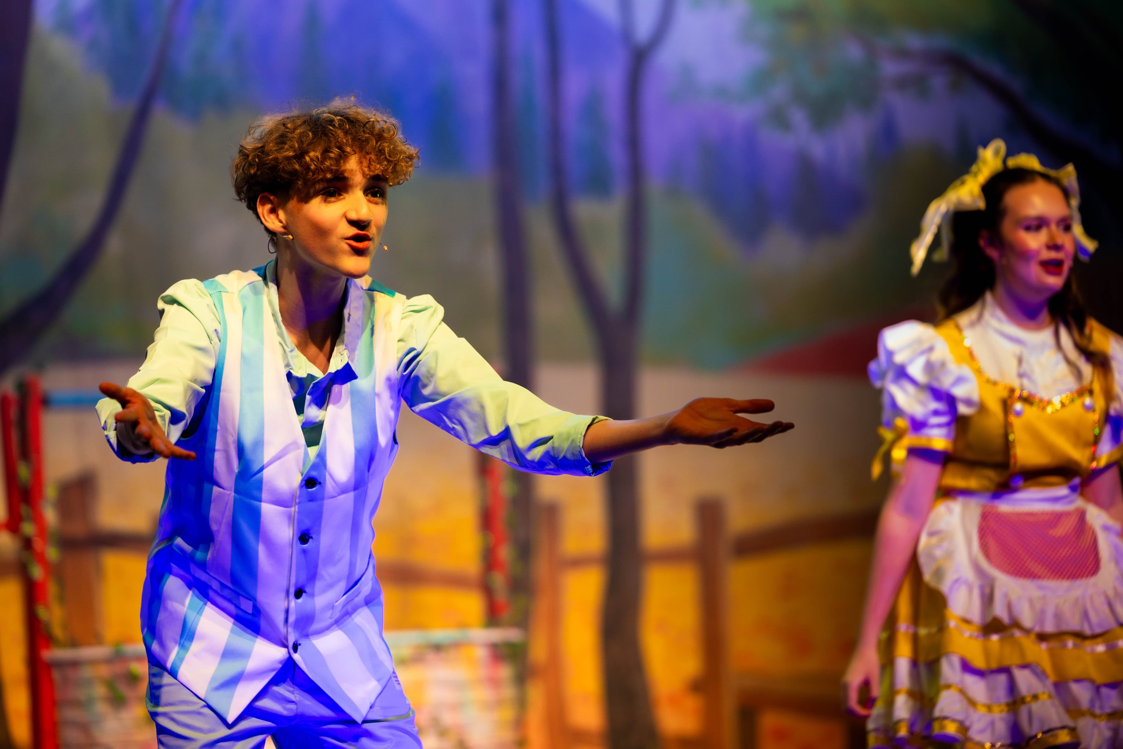 Hansel gestures towards the audience with his arms out. Goldi can be seen in the background.