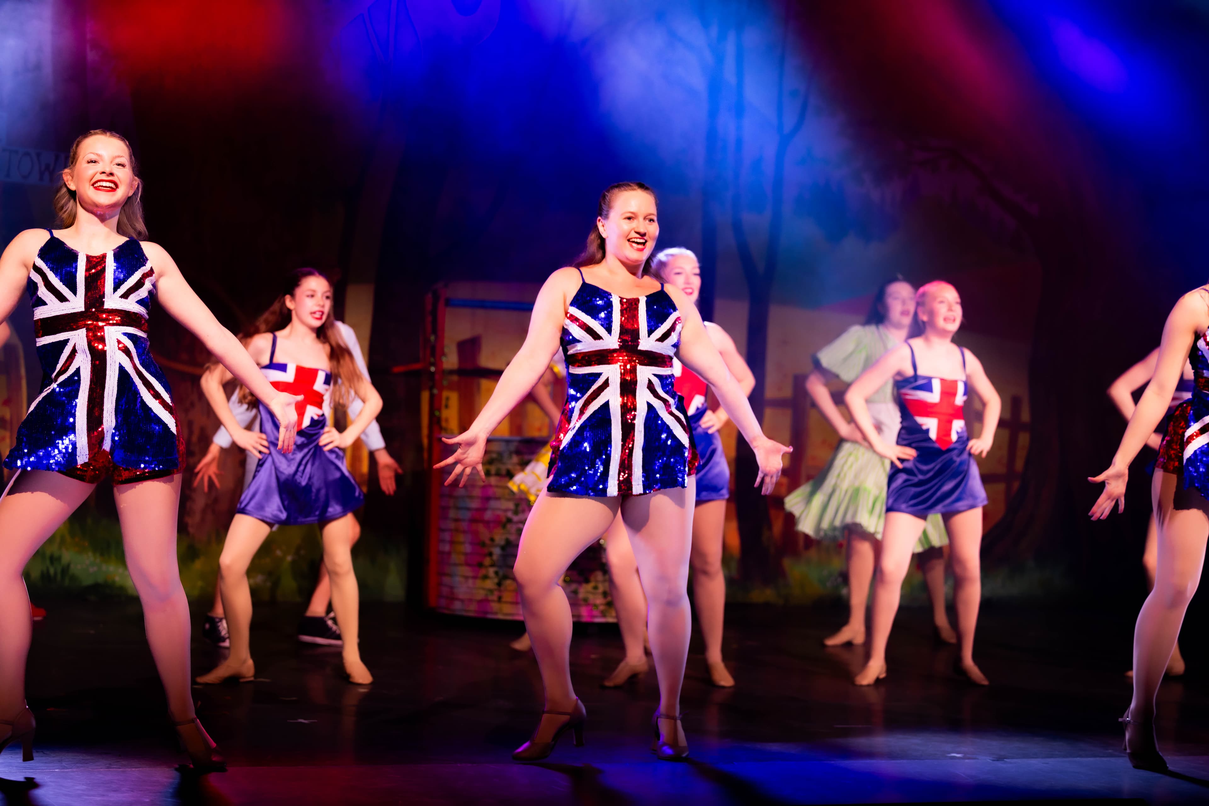 The Principal Dancers dance in sparkly Union Flag dresses. They are lit from behind in red, white, and blue. The rest of the cast dance behind them.