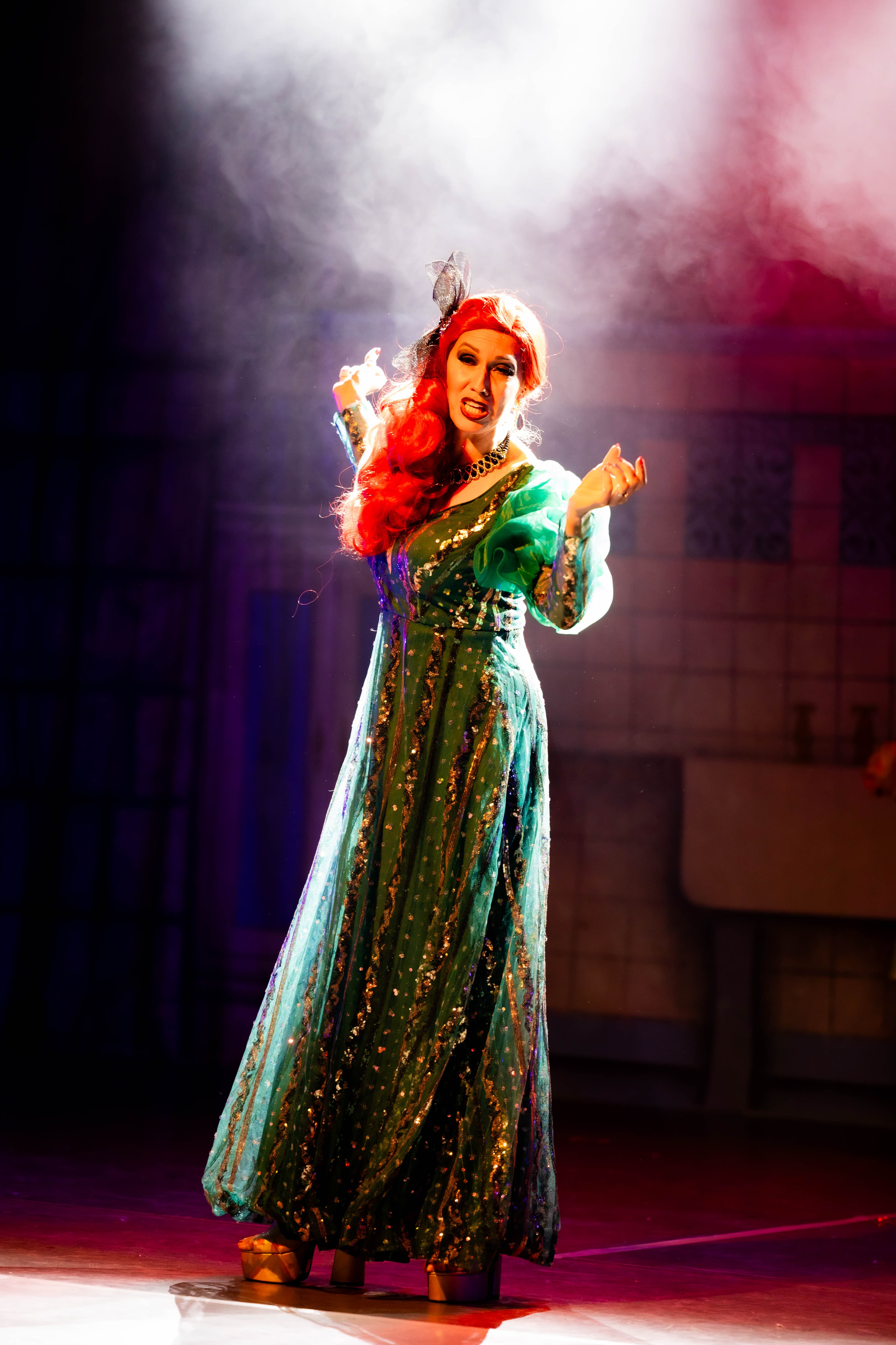 The Wicked Stepmother poses in her green ornate dress in a cloud of spoke. She is lit from above in a white spotlight.