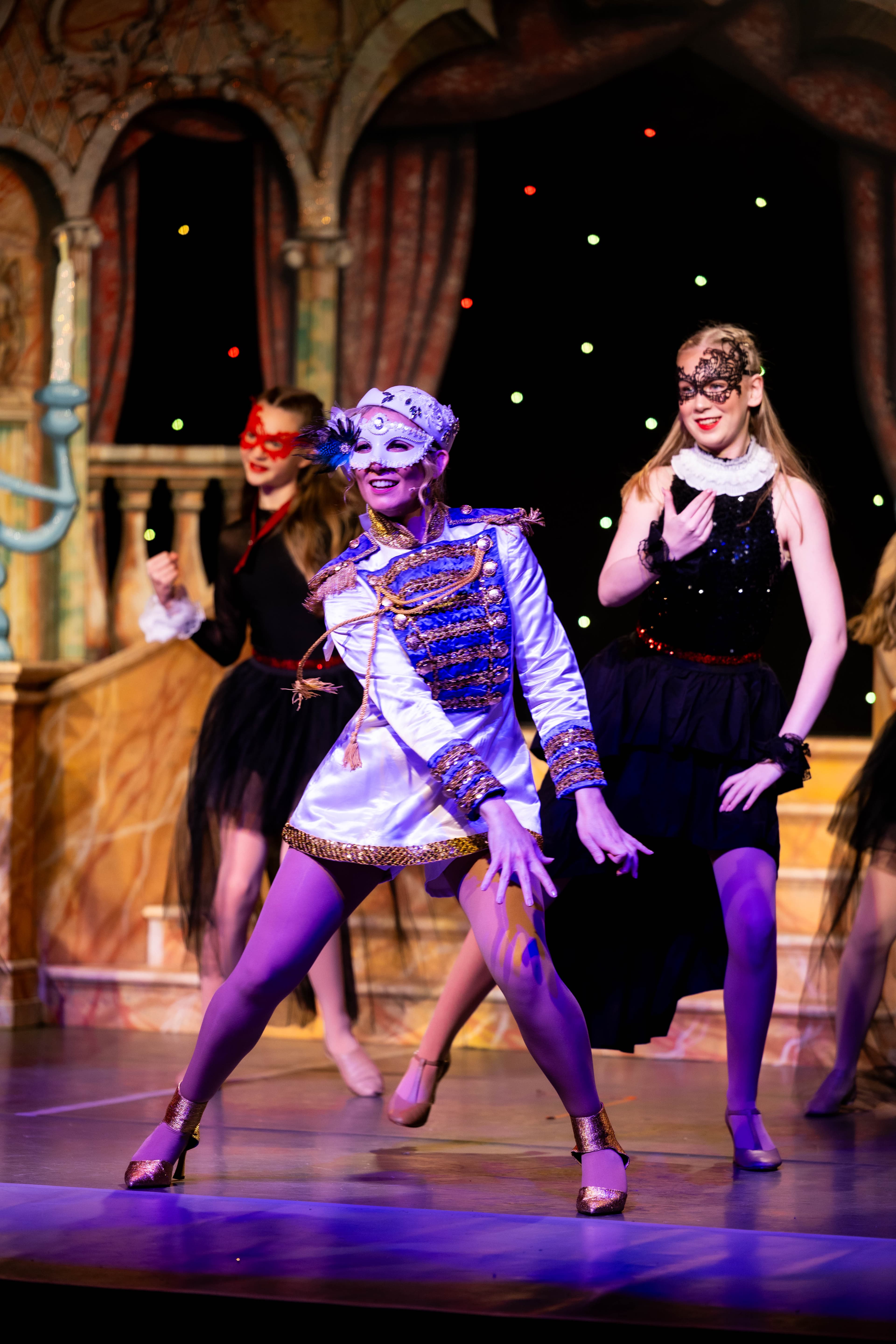 Prince Charming dances during the masquerade ball. He wears a white and blue costume, with a white mask. Dancers in black masquerade dance behind. Stars sparkle behind them.