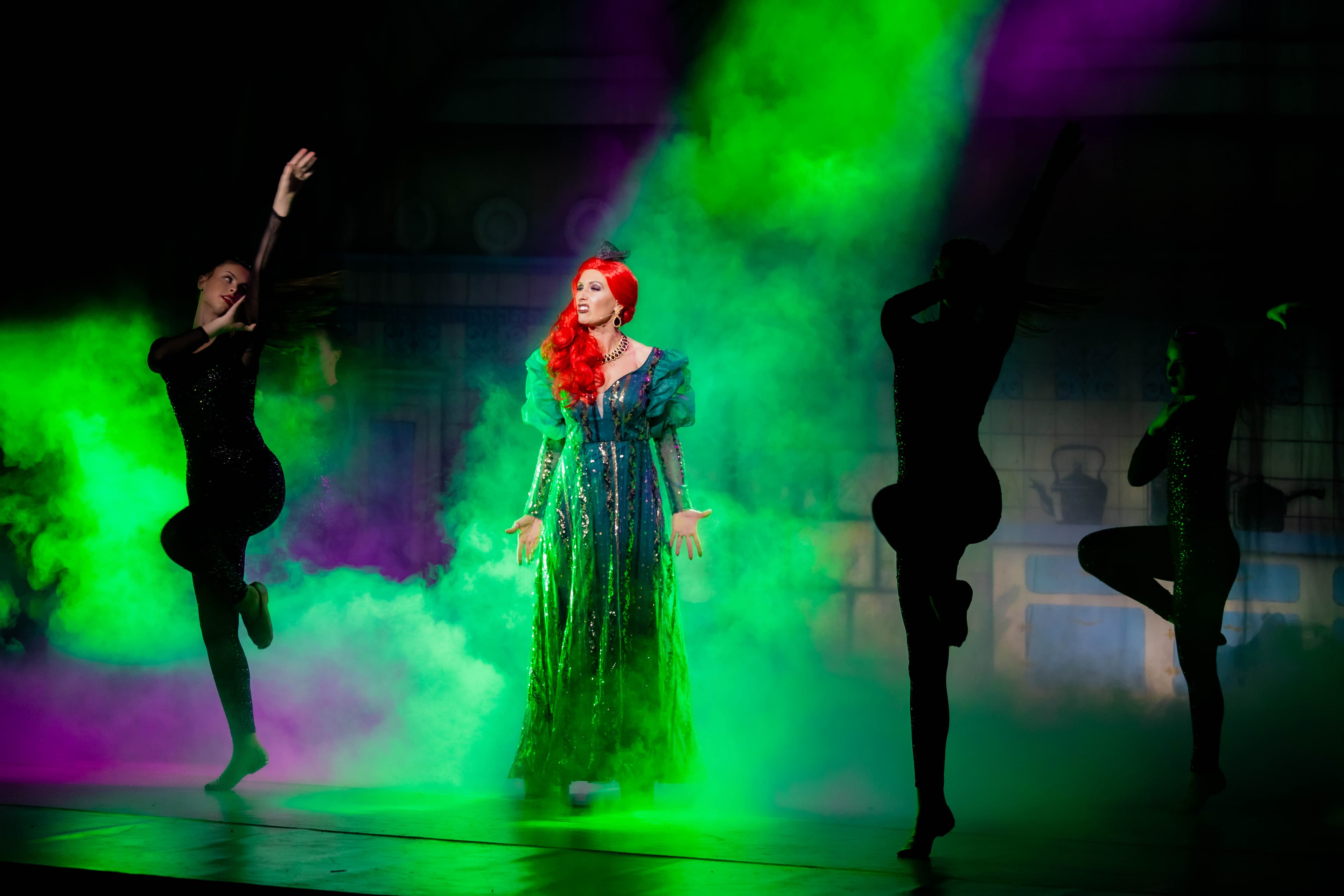 The Wicked Stepmother is pictured posing while singing a song. She stands in a sharp green shaft of light and a cloud of smoke. Silhoetted dancers dance around her.