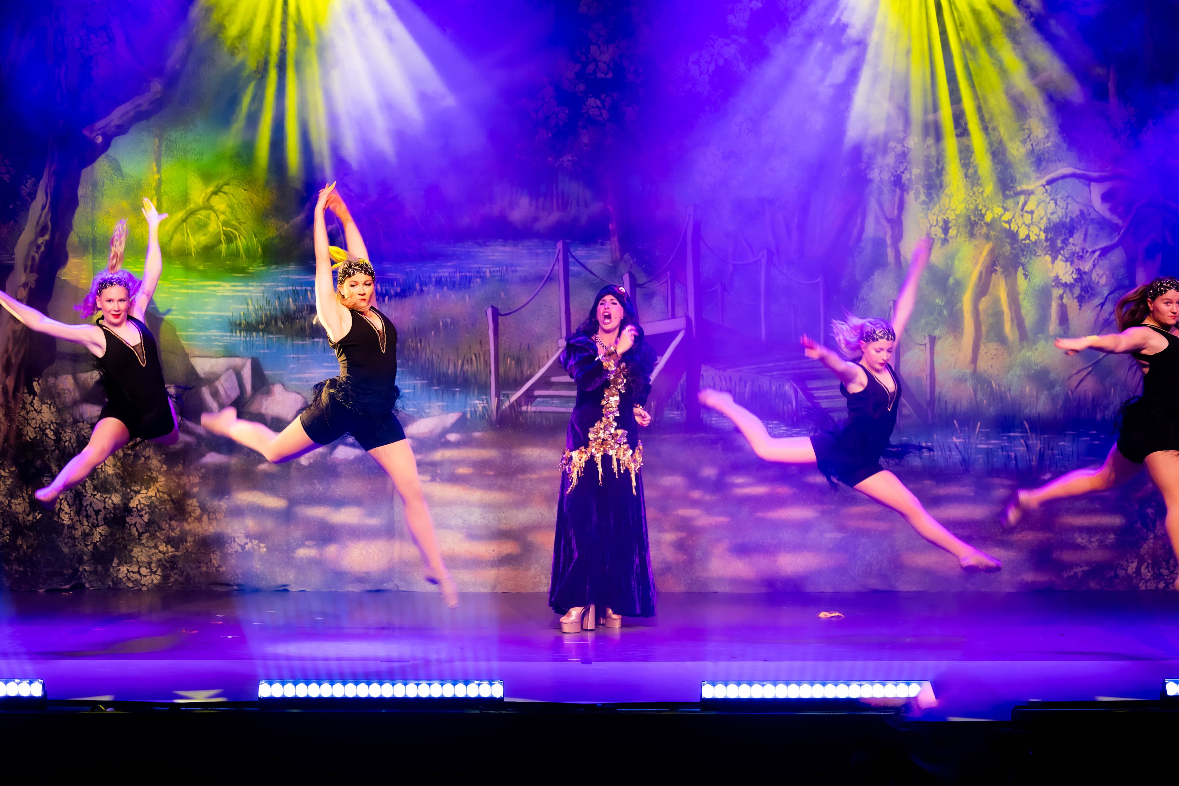 The Principal Dancers are caught mid-leap during a dance while Mother Gothel sings a solo. The stage is lit purple, with yellow spotlights from above.
