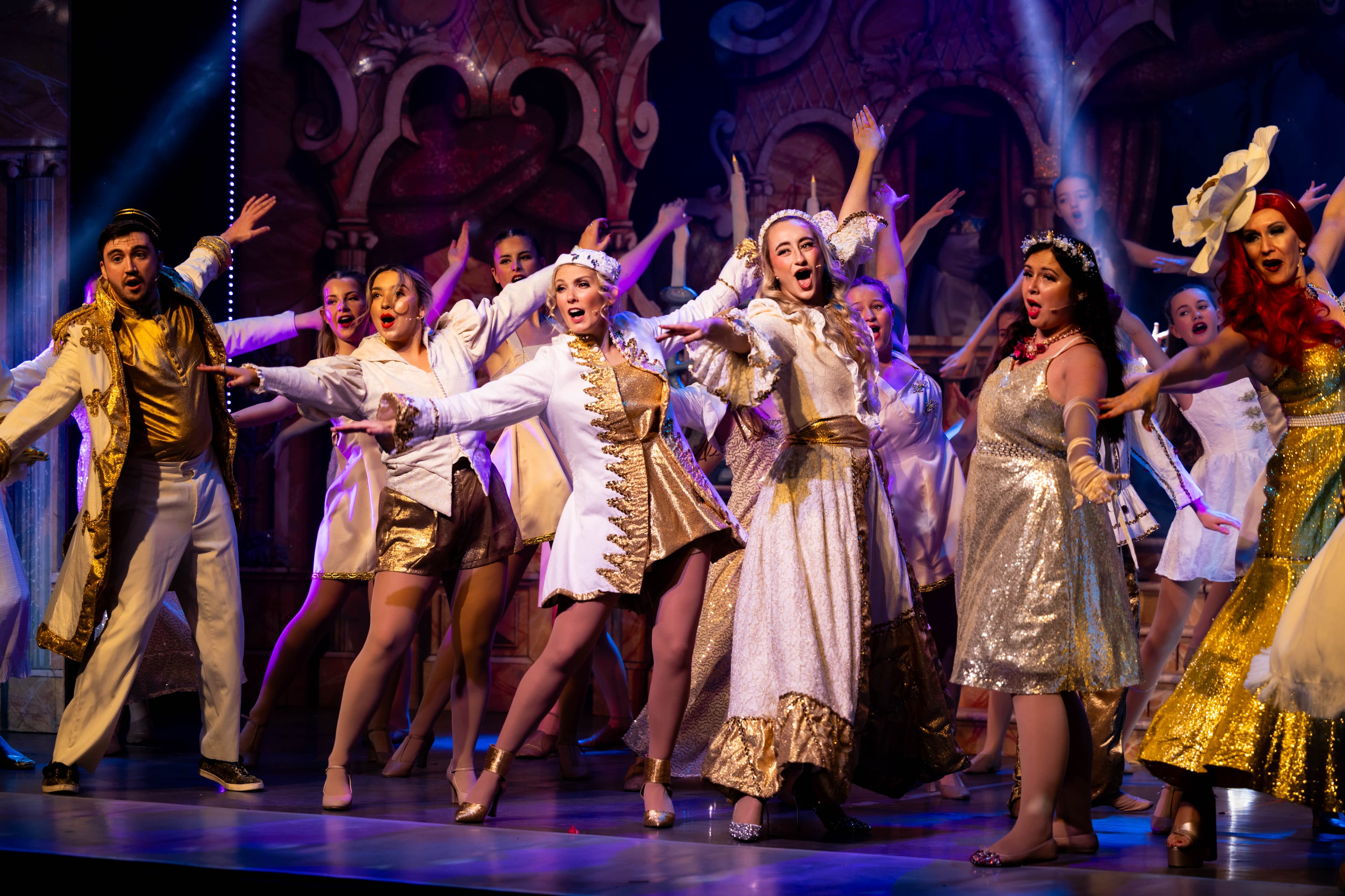 The principal cast dance in ornate gold and while outfits during the finale of the show. They are shown with their arms out, angled down.