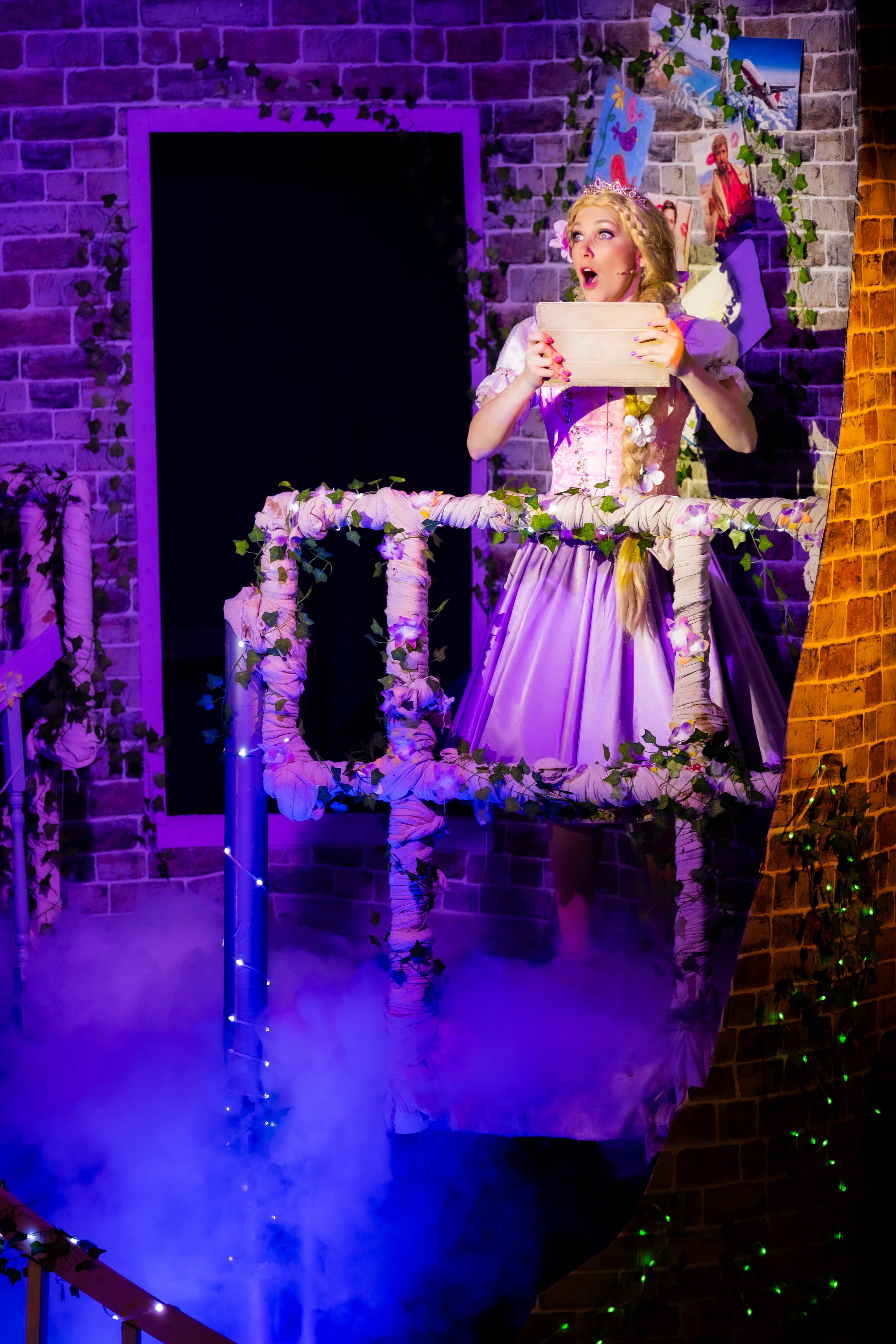Rapunzel is pictured in her tower on her iPad. The tower is lit in purple. She stands atop a glowing glow of blue low fog, and the tower is covered in sparkling LEDs.